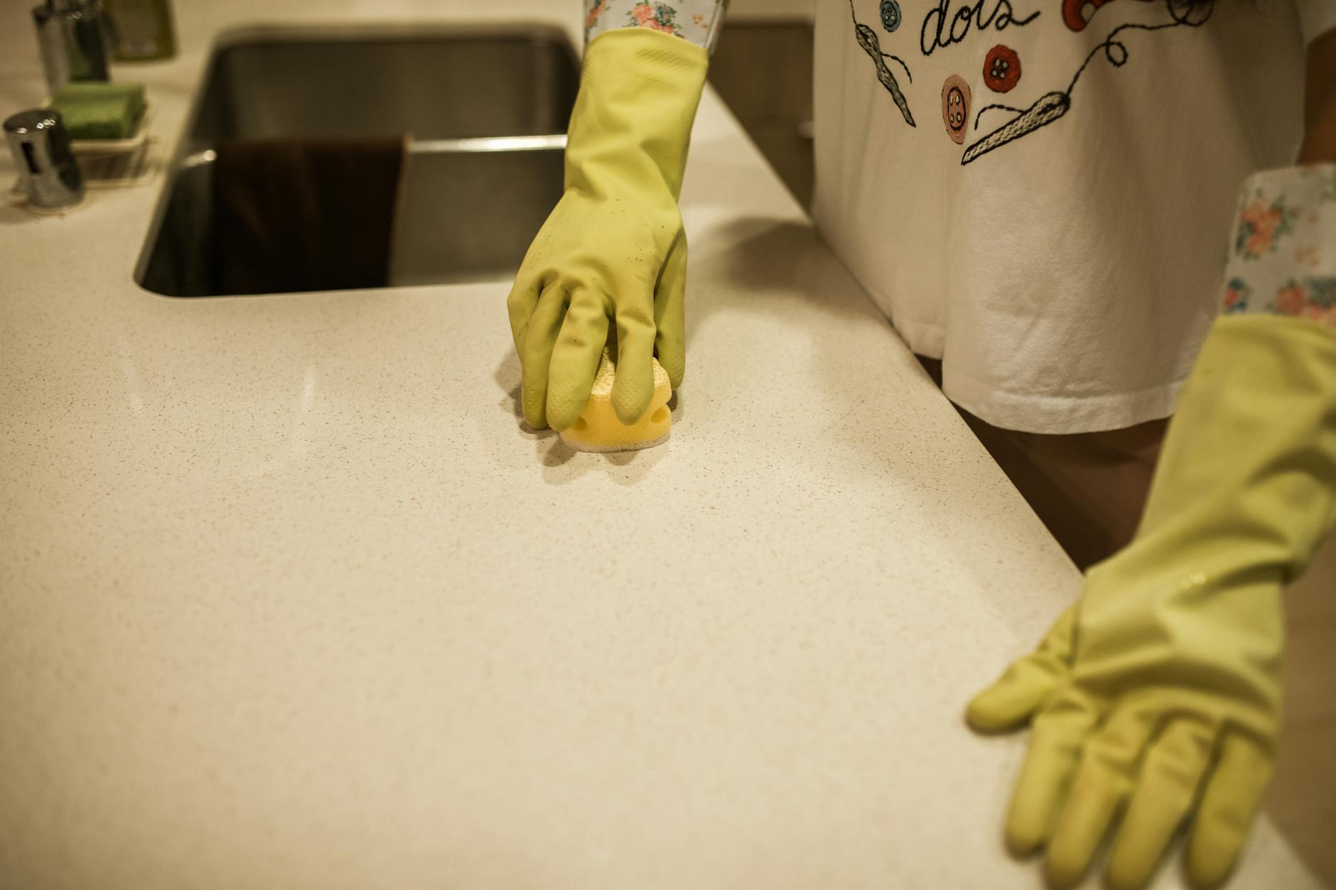 Gloved hand scrubbing a pale kitchen counter with a sponge next to a stainless steel sink