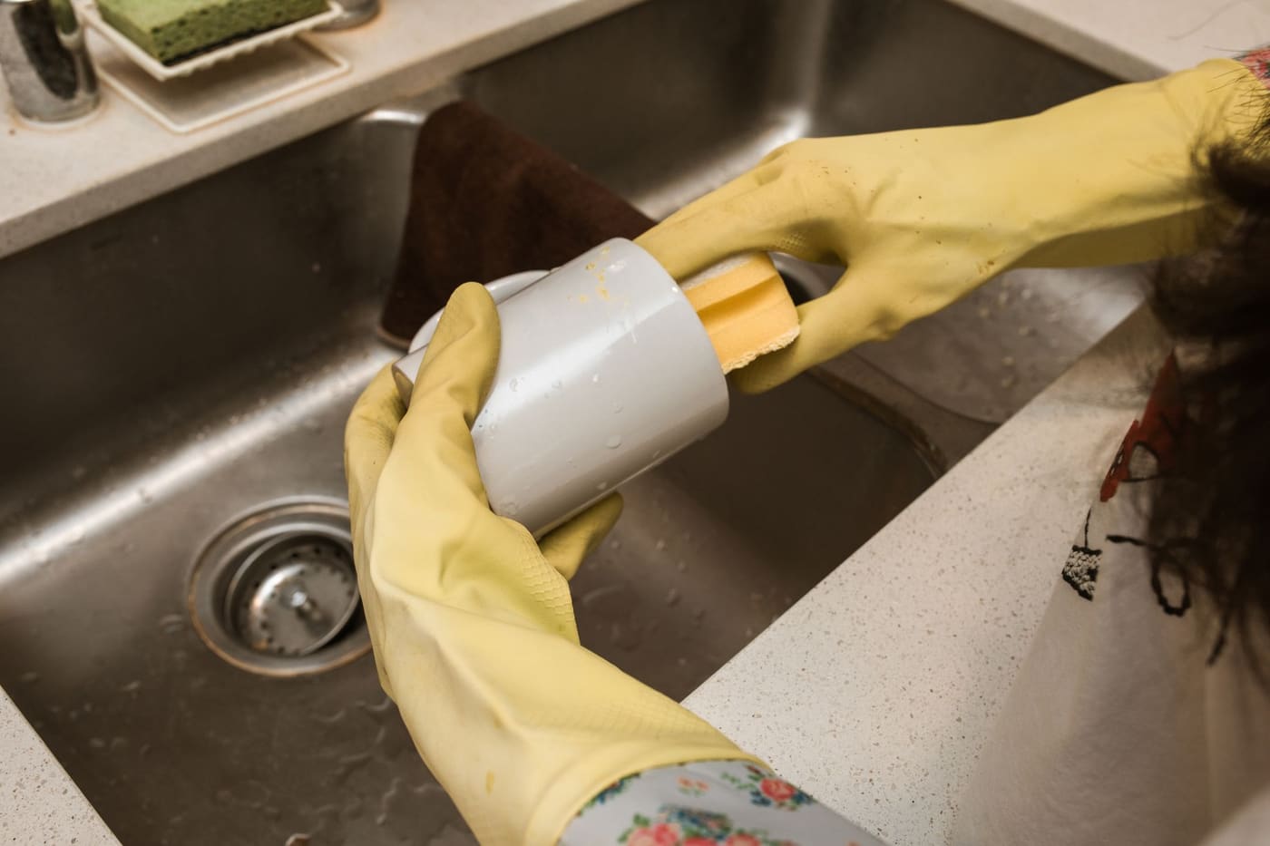 Hands in yellow rubber gloves washing a mug with a sponge in a stainless steel sink