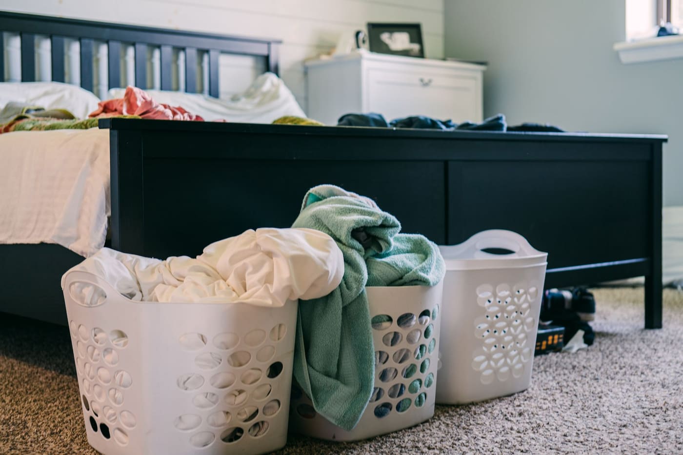 Two plastic laundry baskets of clothes on a bedroom carpet beside a bed