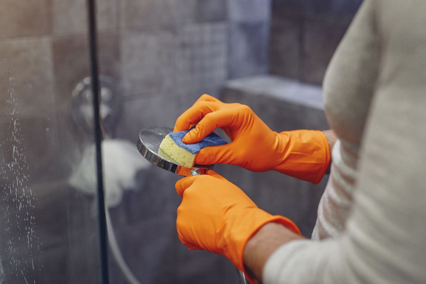 Hands in orange rubber gloves scrubbing a showerhead with a sponge
